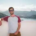 Young man in sunglasses leaning on a balcony with a scenic city view in the background.